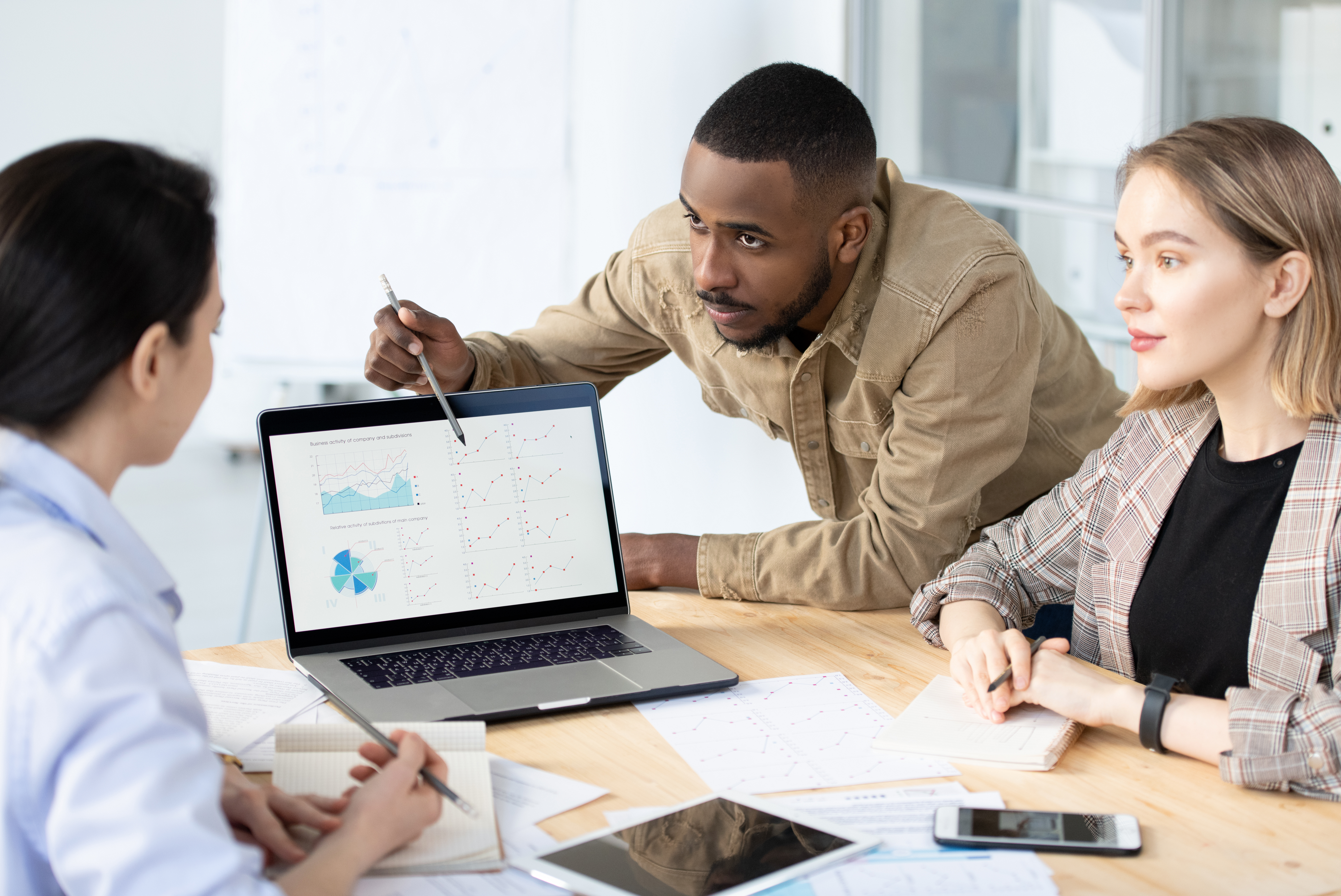 Young confident quant-type bending over a desk showing data to two colleagues.